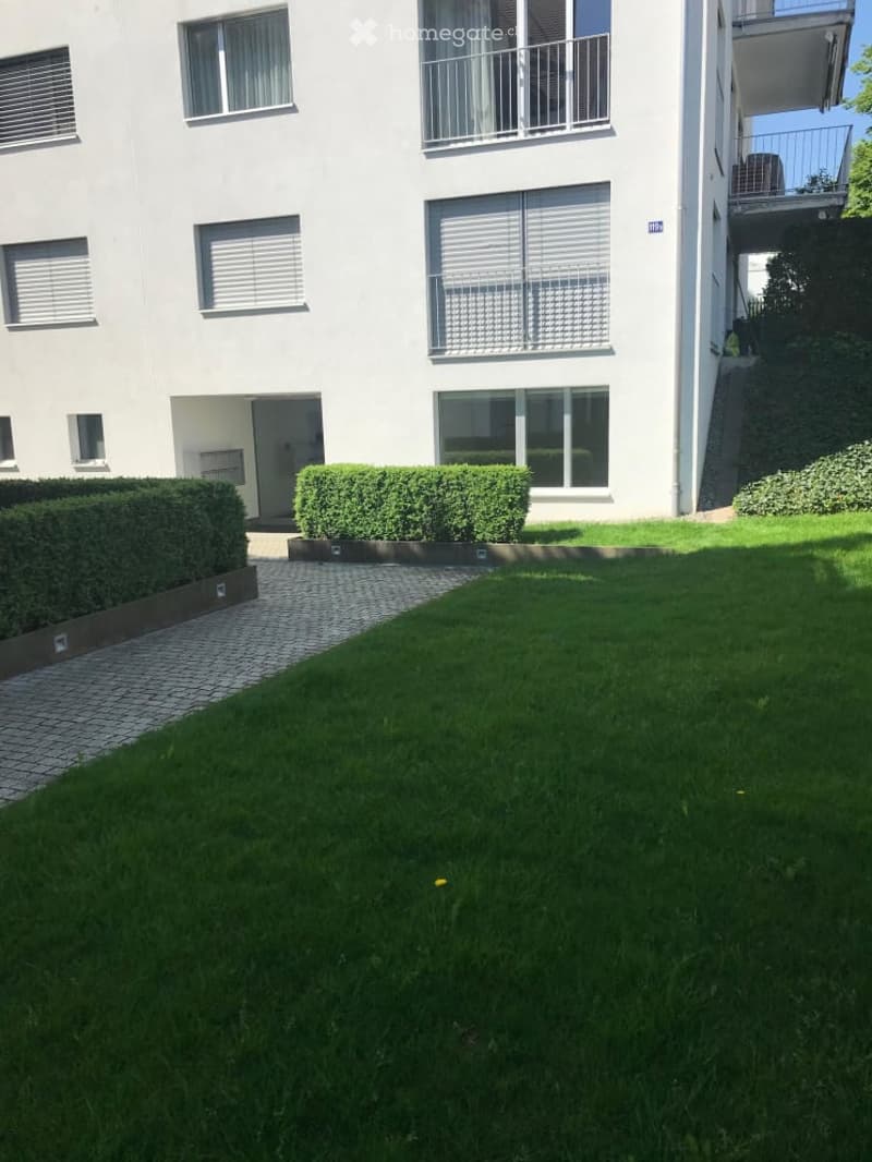 Well-maintained garden area with green lawn, trimmed hedges, and a paved pathway leading to a modern building with balconies.