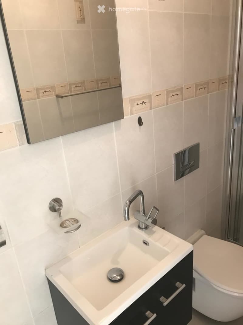 Modern bathroom with a white sink, chrome faucet, mirror, and toilet. The walls are tiled in a light beige color with white accents.