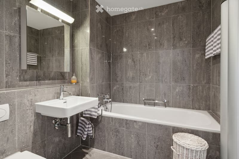 Modern bathroom with dark grey tiled walls, white sink, bathtub, mirror, and striped towels.
