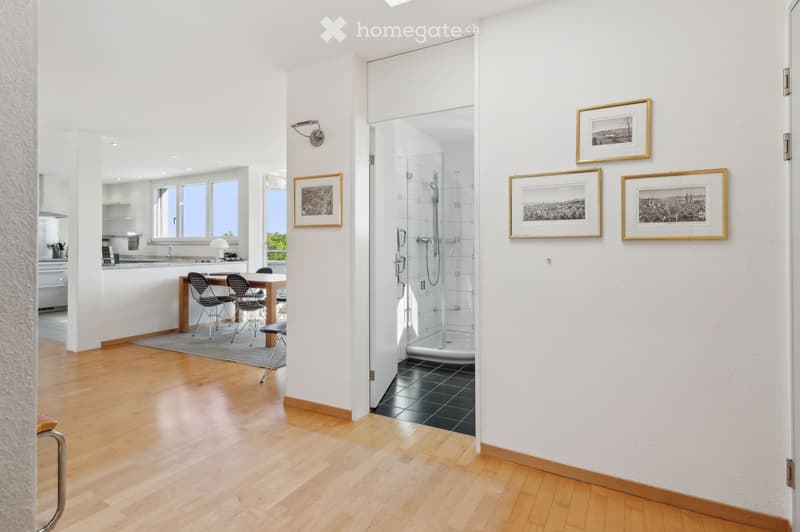 Bright hallway with wooden flooring, leading to a dining area and a modern bathroom with a glass shower enclosure. The walls are white and decorated with framed artwork.