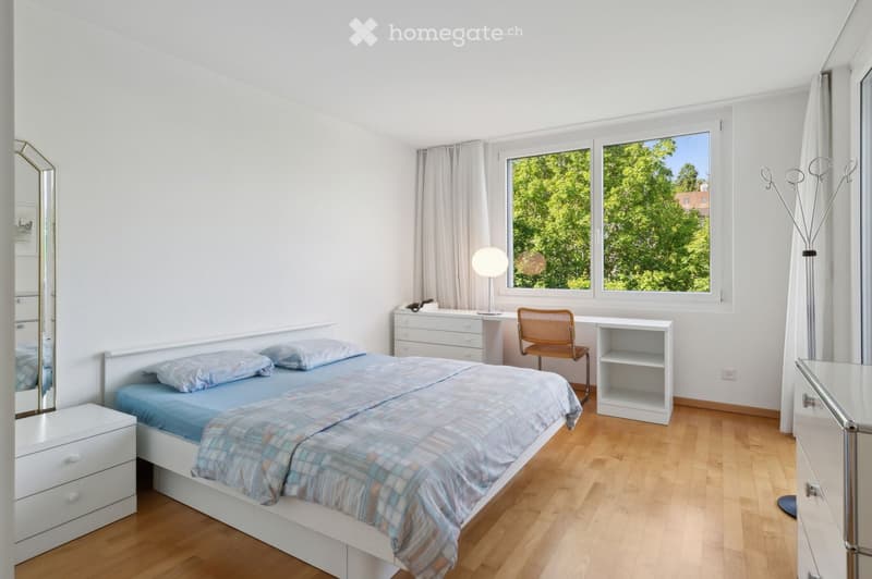 Bright bedroom with wooden flooring, a single bed with blue bedding, a nightstand, a desk with a chair, a lamp, and a large window with curtains overlooking greenery.