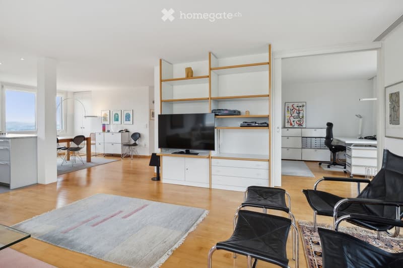 Modern living room with wooden flooring, white walls, and large windows. Features a sofa, chairs, a coffee table, shelves, a television, and a desk area with a chair.