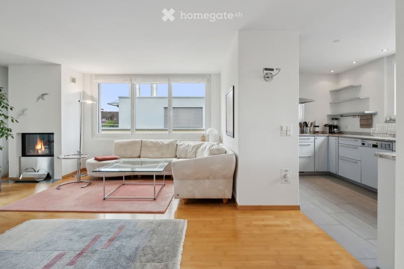 Modern living room with a beige sofa, glass coffee table, fireplace, and an open kitchen area with white cabinets and stainless steel appliances.