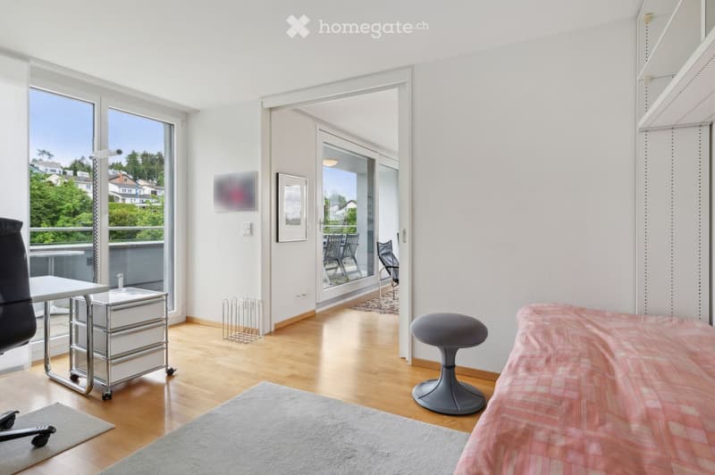 Bright bedroom with wooden flooring, large windows, and a balcony. Features a bed with a pink bedspread, a grey stool, a chair, a small table, and a framed picture on the wall.