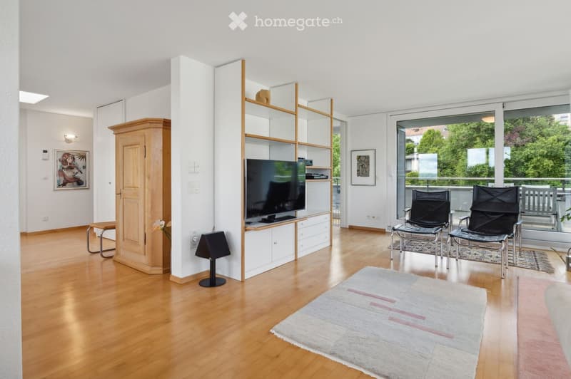 Modern living room with wooden flooring, large windows, and a balcony view. Features include a sofa, chairs, a coffee table, a television, a cabinet, and a floor lamp.
