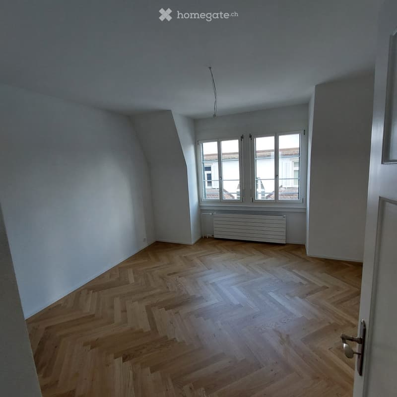Empty room with wooden herringbone flooring, white walls, and large windows with white frames. A radiator is visible beneath the windows.