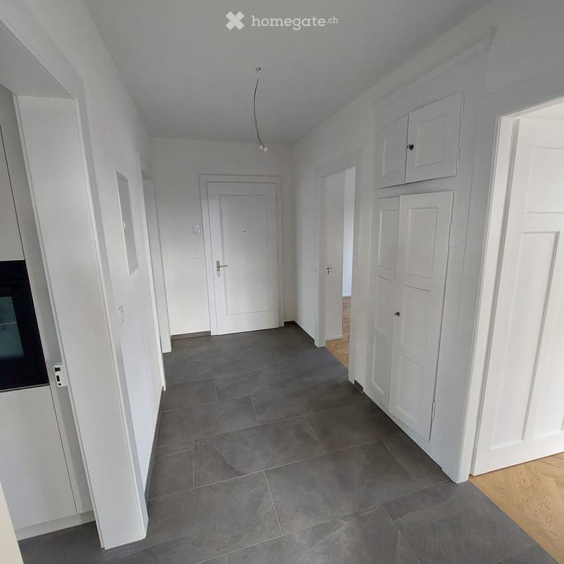 Empty hallway with grey tiled floor, white walls, and several white doors leading to other rooms. The hallway has a slanted ceiling and a small light fixture hanging from it.