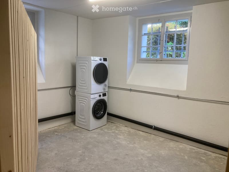 A small, unfurnished room with a washing machine and dryer stacked together, a window, and a concrete floor.