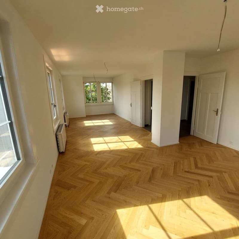 Empty room with light wooden herringbone flooring, white walls, and large windows allowing natural light to fill the space. Radiators are visible along the walls, and there are several open doors leading to other areas.