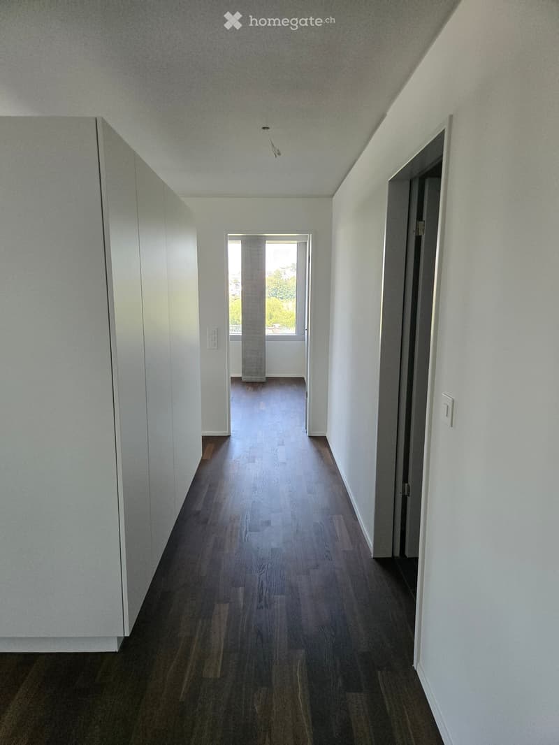 Empty hallway with wooden flooring, white walls, and a ceiling light fixture. A doorway leads to a room with a window and a view of greenery outside.