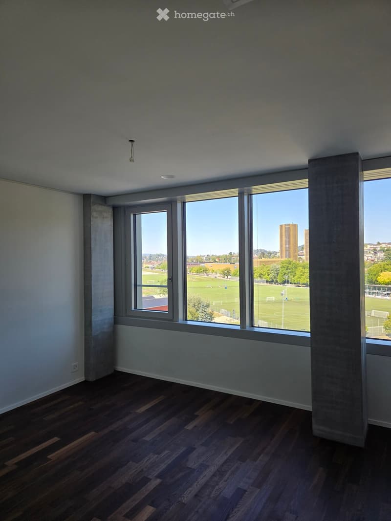 Empty room with wooden flooring, large windows overlooking a sports field, and modern interior design.