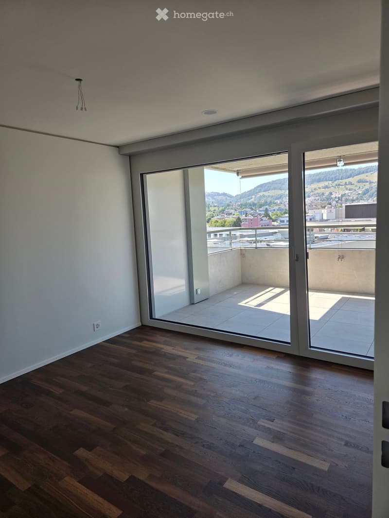 Empty room with wooden flooring, large sliding glass doors leading to a balcony with a view of the city and surrounding landscape.
