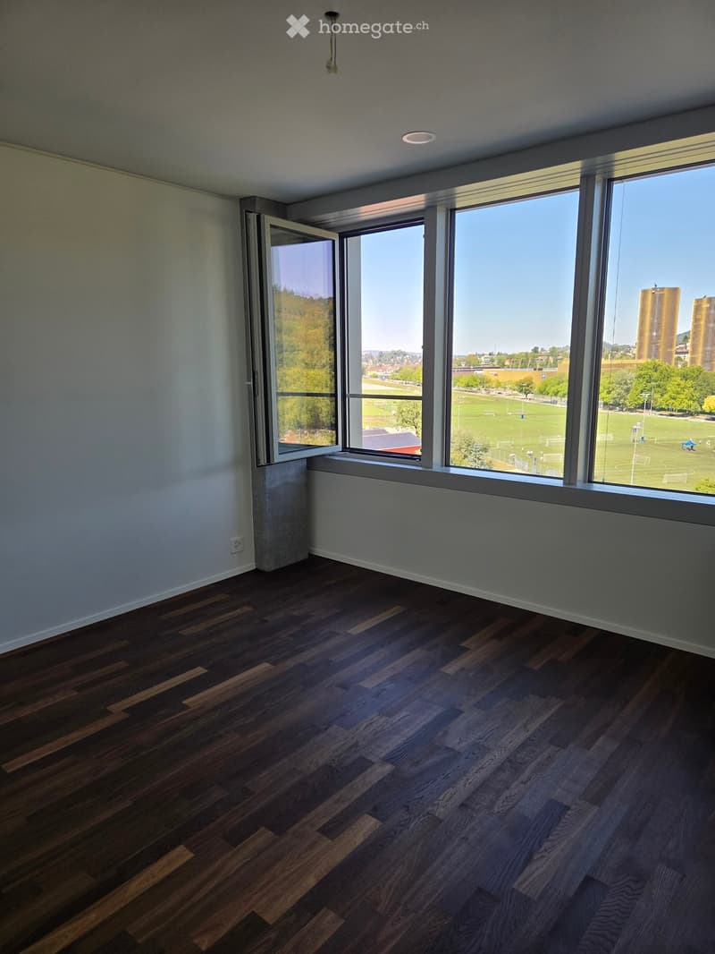 Empty room with dark wooden flooring, large windows offering a view of a green field and distant buildings, and white walls.