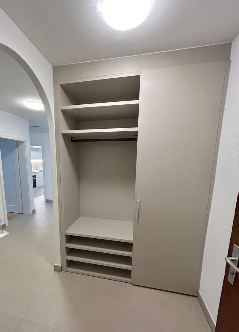Empty closet with built-in shelves and a hanging rod, located in a hallway with tiled flooring and white walls.