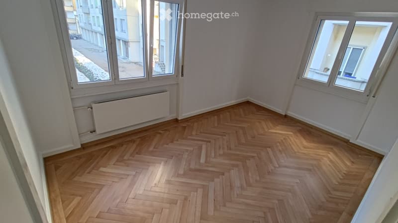 Empty room with herringbone wooden flooring, large windows, radiator, and white walls.
