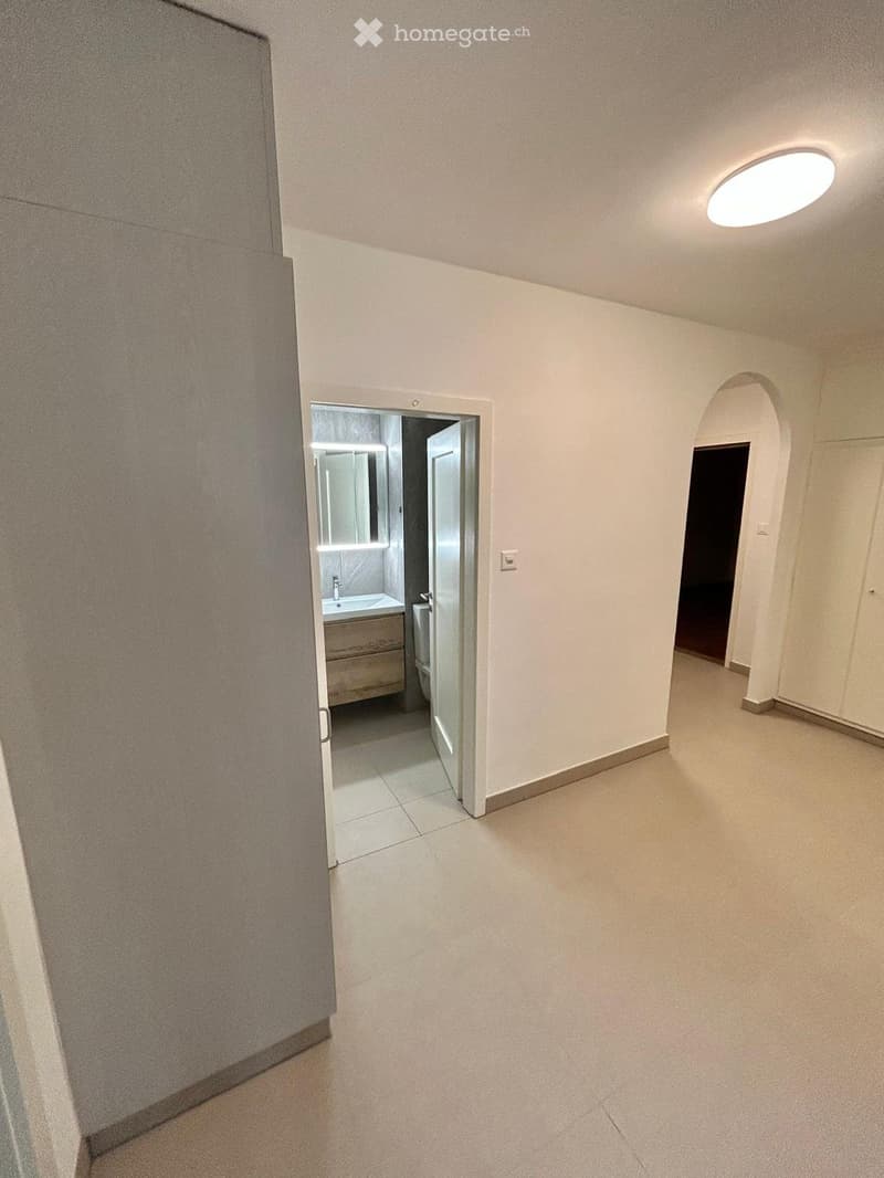Empty hallway with white walls, tiled floor, and a round ceiling light. An open door leads to a small bathroom with a sink, mirror, and cabinet.