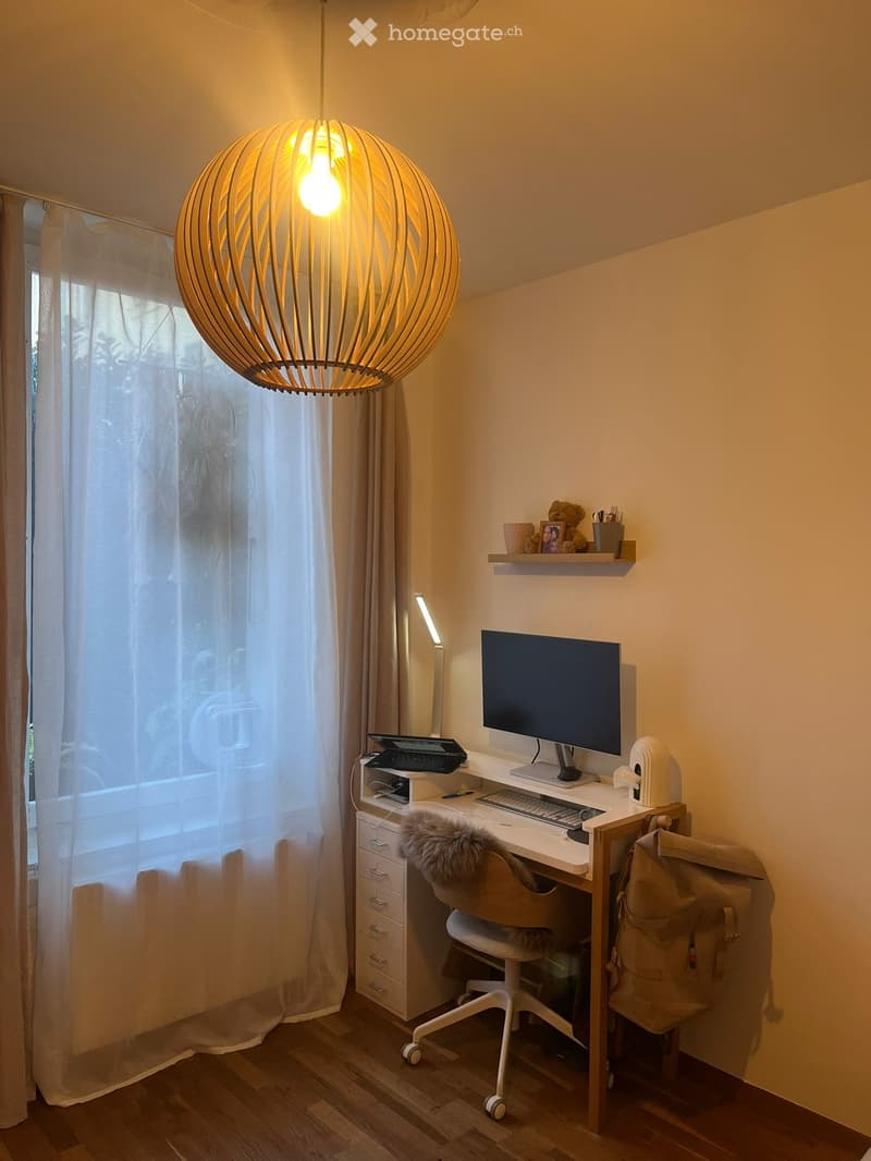 A modern, well-lit workspace with a wooden desk, ergonomic chair, laptop, monitor, keyboard, mouse, and a decorative pendant light. The room features large windows with sheer curtains and a small shelf with decorative items.
