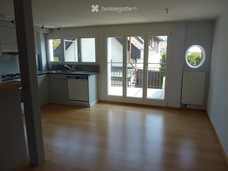 Empty room with wooden flooring, large windows, and a balcony door leading to a balcony. Kitchen area visible with sink, stove, and dishwasher.