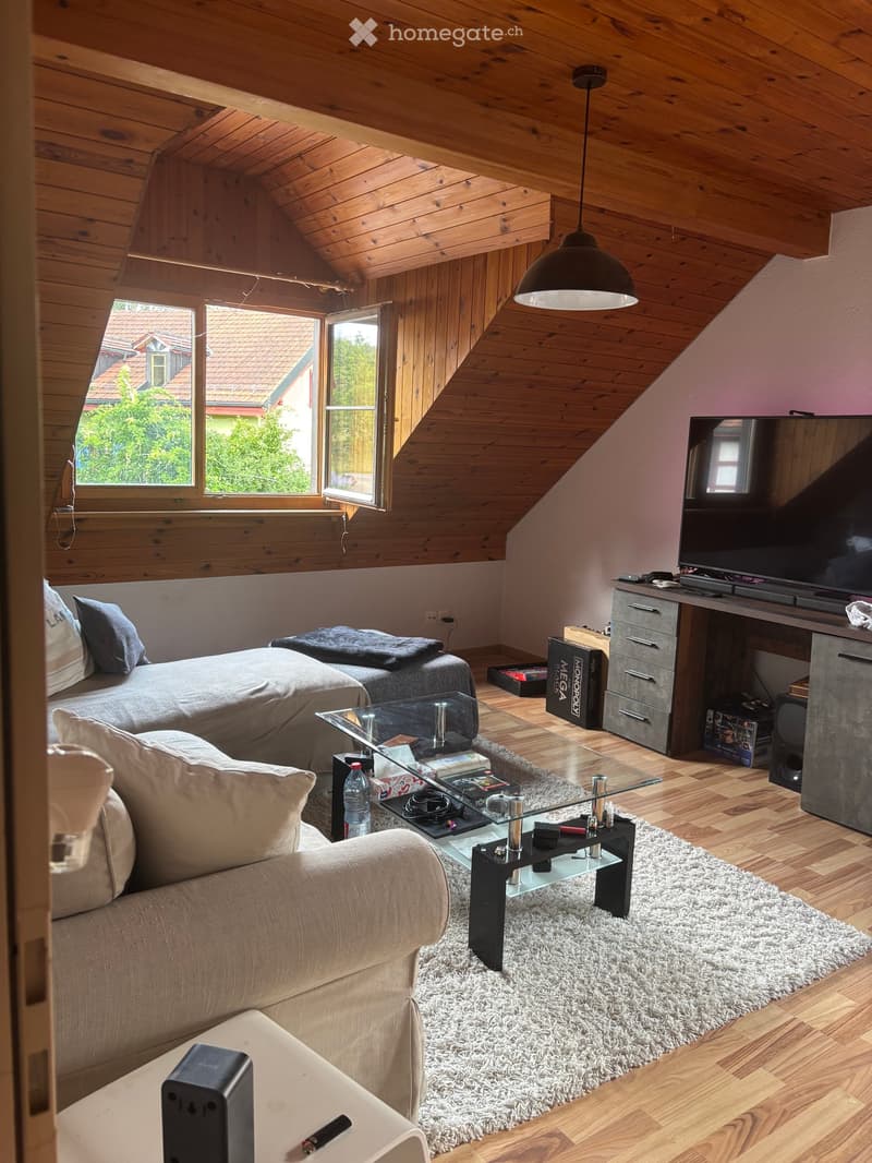 A cozy attic room with wooden beams, a large window, a sofa, a coffee table, a bed, a television, and a desk.