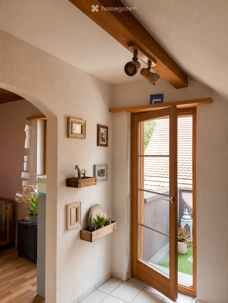 A narrow hallway with wooden beams on the ceiling, white walls, and a wooden door leading to an outdoor area. Shelves with decorative items and framed pictures are mounted on the wall. The floor is a combination of wooden planks and white tiles.