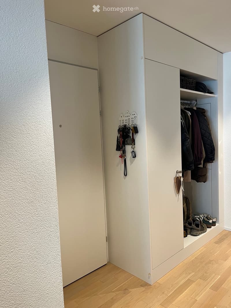 A modern hallway with a built-in closet featuring white cabinetry, hanging coats, shoes, and keys on a wall-mounted key holder.