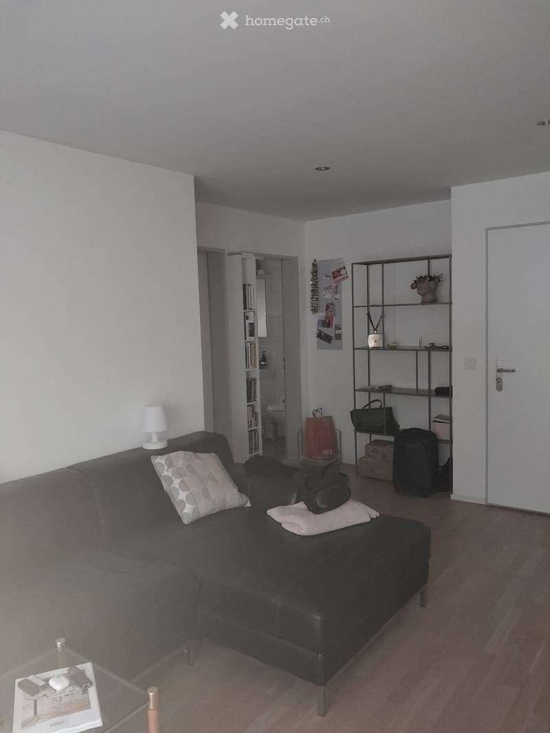 A modern living room with a grey sofa, a small table, a shelf, and a lamp. The room has light-colored walls and wooden flooring.