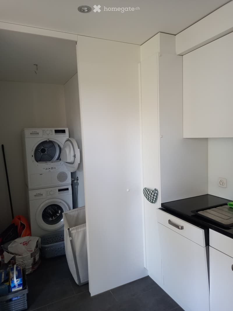 A compact bathroom with a stacked washing machine and dryer, white cabinets, a black countertop, and a small trash bin.