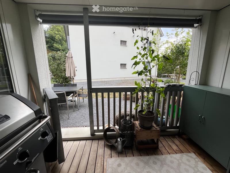 Balcony with wooden decking, grill, small table and chairs, potted plant, and a view of a garden area.