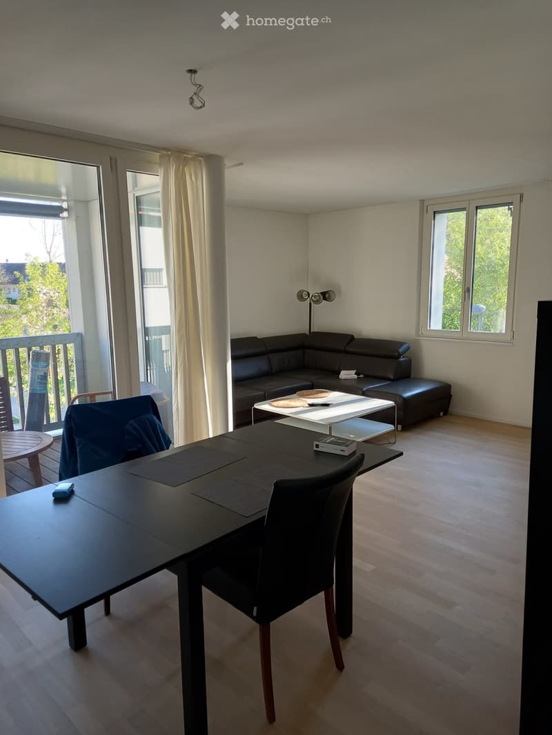 Modern living room with a black sectional sofa, coffee table, dining table, and chairs. Large windows and a balcony are visible.