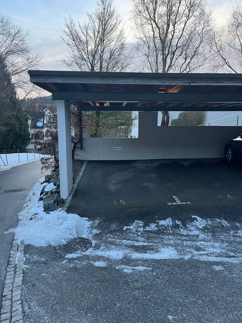 covered parking space with a car parked in it, concrete wall, white markings on the floor, and an outdoor area covered with snow
