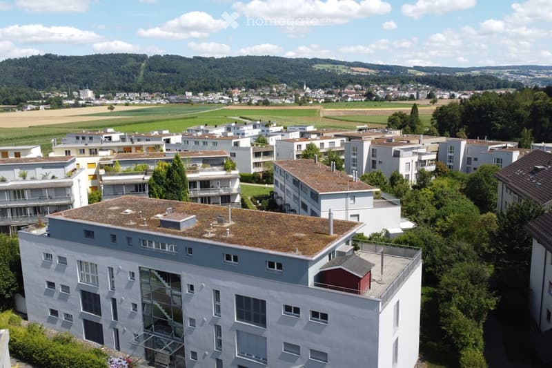 Aerial view of a modern multi-story residential building with a green roof, surrounded by other buildings and greenery, with a scenic countryside backdrop.