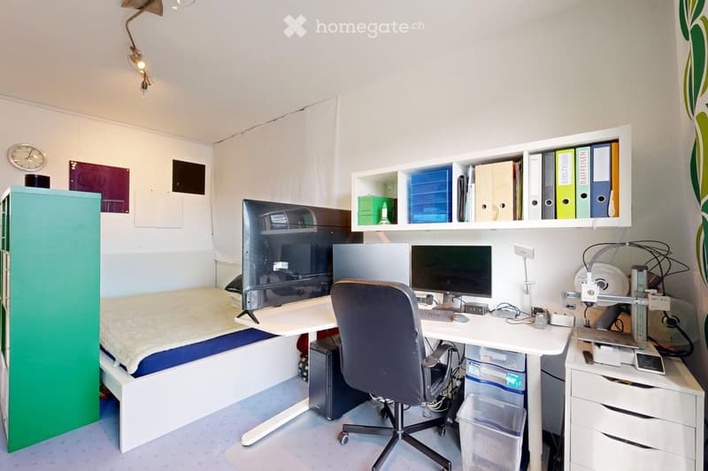 A compact, multi-functional room featuring a bed, a desk with a computer setup, shelves with binders and storage boxes, and a wall-mounted lamp. The room includes a chair, a fan, and a small trash bin.