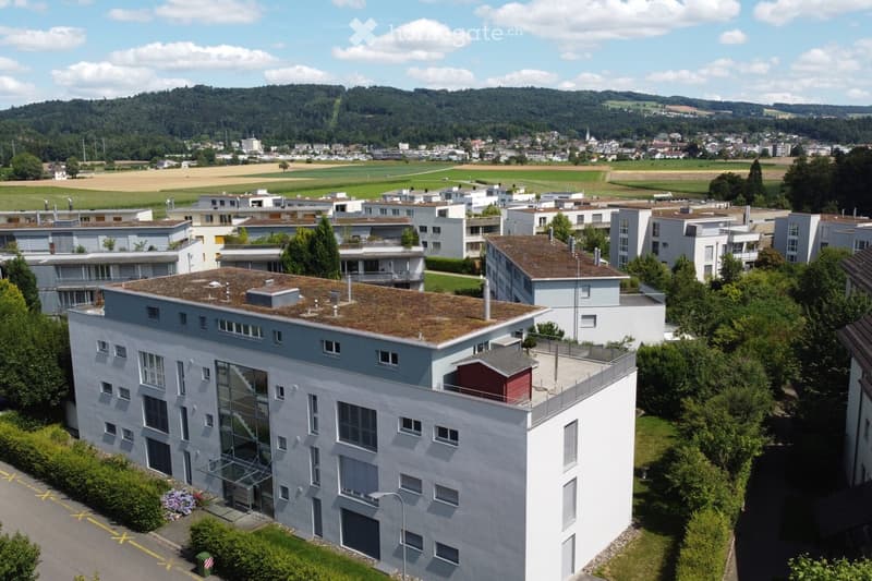 A modern multi-story building with a green roof, surrounded by trees and other residential structures in a suburban area with a scenic backdrop of hills and fields.