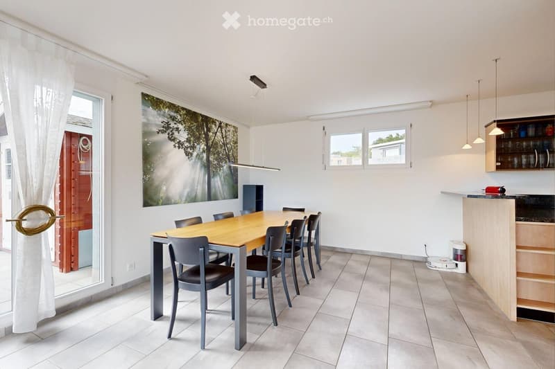 Bright dining room with a wooden dining table surrounded by six chairs, a large window with white curtains, and a cabinet with glass doors.