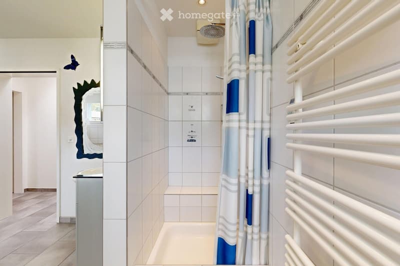 Modern bathroom with white tiled walls, a shower area with a blue and white striped curtain, a radiator, and a decorative mirror with a butterfly design.