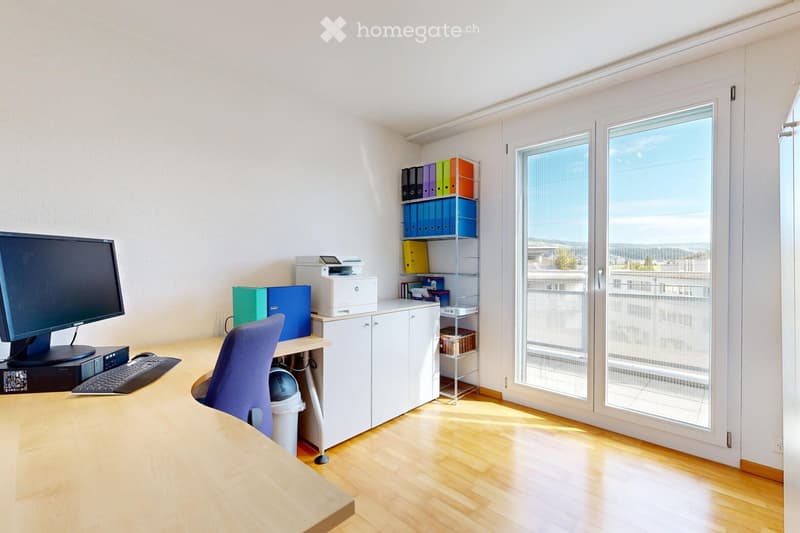 Bright office space with wooden flooring, large windows, desk with computer setup, chair, cabinet, and shelving unit with binders.