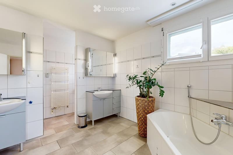 Modern bathroom with white tiles, two sinks, a bathtub, a large mirror, a radiator, and a potted plant near the window.