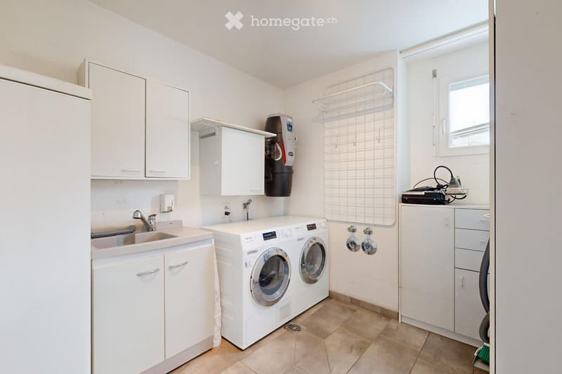 A modern bathroom with a sink, washing machine, dryer, and water softener. The space features white cabinets, a tiled floor, and a laundry rack with a mirror above it.