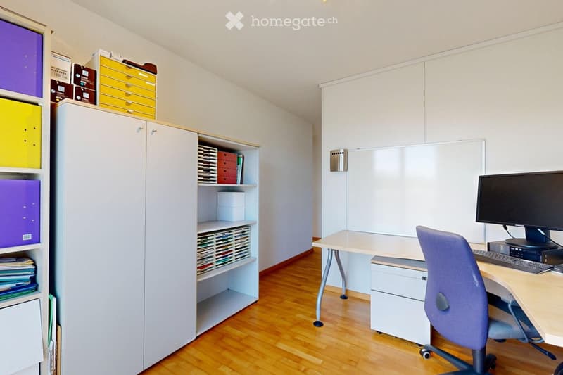Modern office space with a wooden floor, white walls, a desk with a computer monitor, keyboard, and a rolling chair, along with a large cabinet and shelves filled with storage boxes and files.