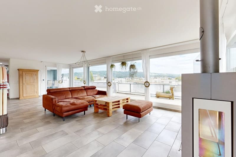 Modern living room with large windows offering a scenic view, featuring a brown leather sofa, wooden coffee table, ottoman, and hanging plants.