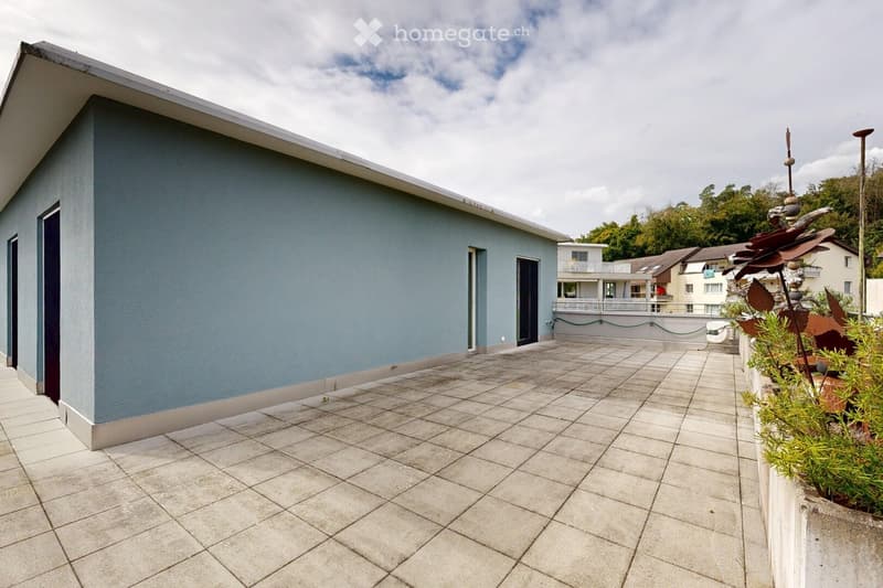 A modern, tiled balcony with a grey wall, small garden area, and a view of neighboring buildings and greenery.