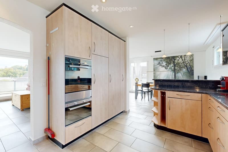 Modern kitchen with light wood cabinets, stainless steel oven, dark countertop, and a small dining area with a table and chairs.