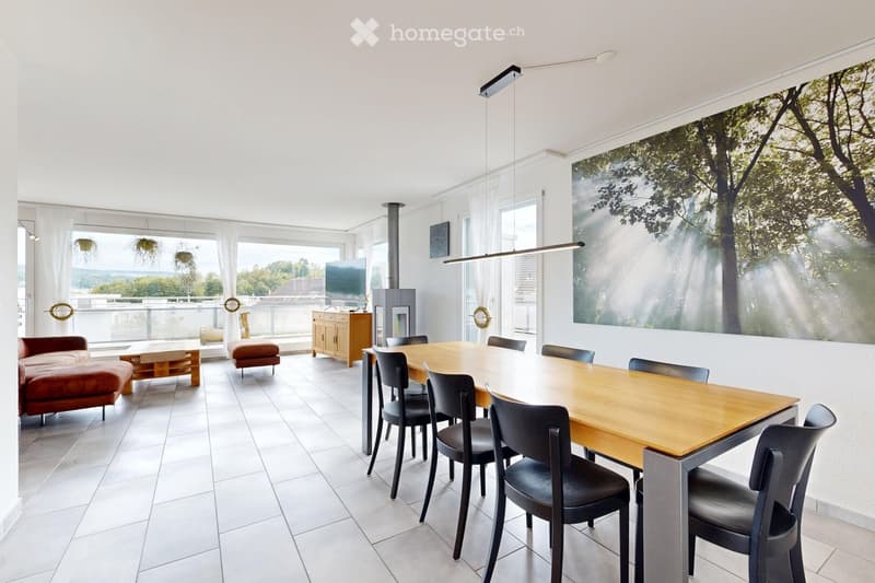 Bright living and dining area with white walls, large windows, and a balcony view. Features a red sofa, ottoman, coffee table, dining table with chairs, a cabinet, and a television. A fireplace is present, and modern pendant lights hang from the ceiling.