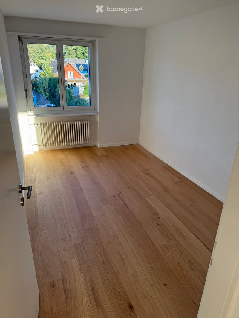 Empty room with light wooden flooring, white walls, a window with a view of the street, and a radiator below the window.