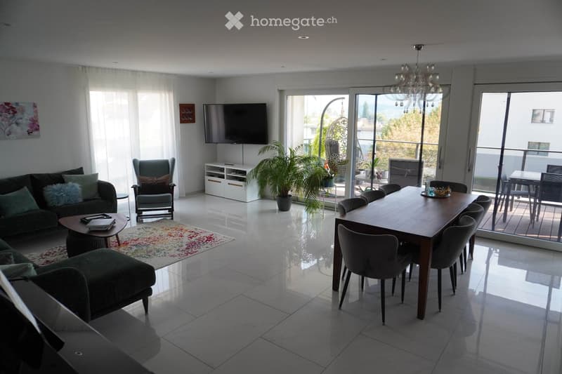 Modern living and dining area with a sofa, ottoman, armchair, dining table, chairs, television, chandelier, and plants. Large windows and sliding glass doors lead to a balcony.