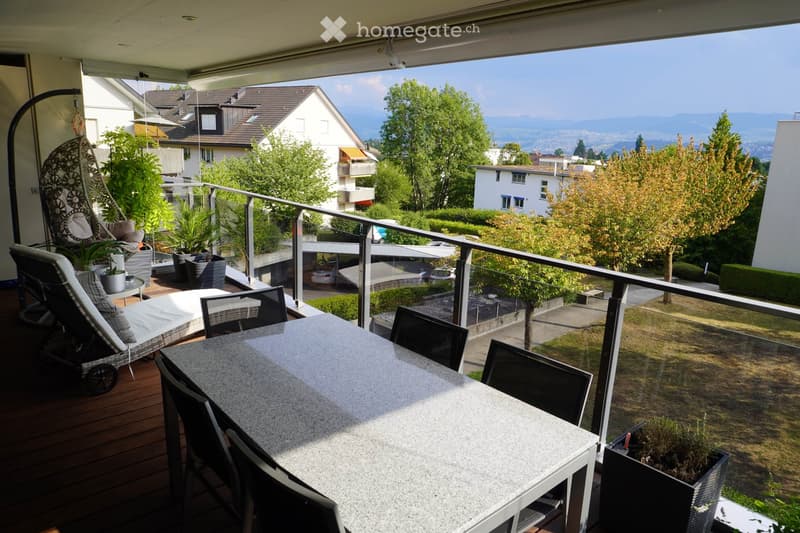Balcony with a table, chairs, wicker hanging chair, and potted plants, offering a scenic view of the surrounding area and mountains.