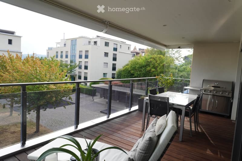 Balcony with wooden decking, glass railing, outdoor furniture including a sofa, table, chairs, and a grill, with a potted plant and scenic city view.