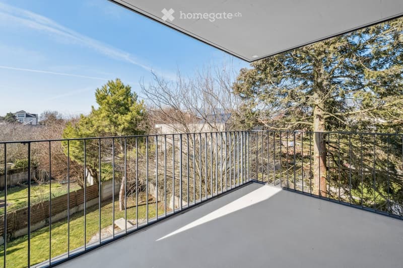 Modern balcony with glass railings, offering a view of a green garden and trees.