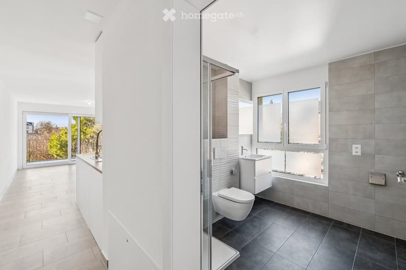 Modern bathroom with white walls, grey tiles, a toilet, sink, bathtub, and a large window offering a view of greenery.
