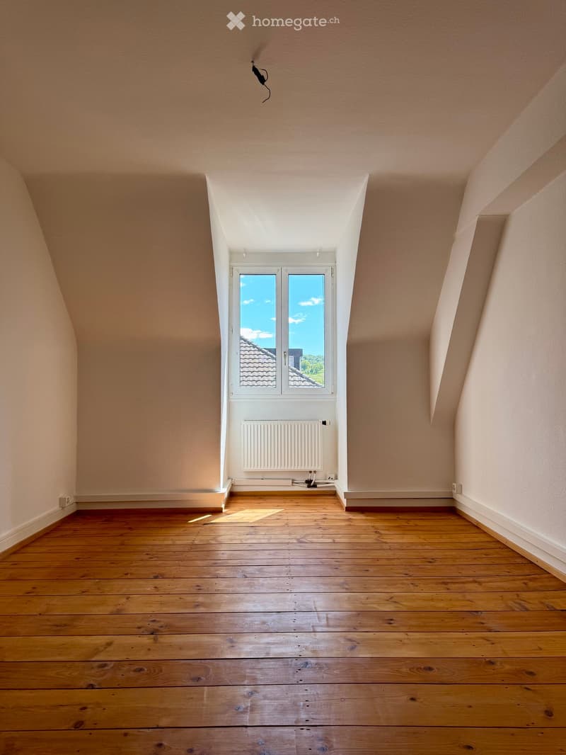 Empty attic room with wooden flooring, slanted ceilings, and a large window offering a view of the roof and sky.